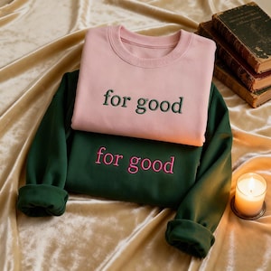 May include: Two sweatshirts, one pink and one dark green, are stacked on top of each other. The pink sweatshirt has the words "for good" embroidered in green, while the green sweatshirt has the same text embroidered in pink. A lit candle and old books are in the background.