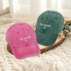 May include: Two baseball caps, one pink and one green, are displayed. Both caps have the phrase "for good" embroidered in white, with a heart symbol. The caps are made of a soft, textured fabric.