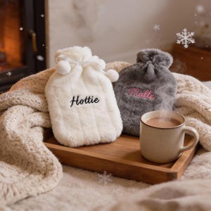 Può includere: Due borse dell'acqua calda in peluche, una bianca con "Hottie" ricamato, l'altra grigia con "Malte". Una tazza di bevanda calda è su un vassoio di legno con le borse. La scena è accogliente.