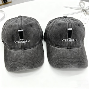 May include: Two gray baseball caps with a distressed finish. Each cap features a black graphic of a glass with a white top and the text "VITAMIN G" in white. The caps have a curved brim and a button on top.