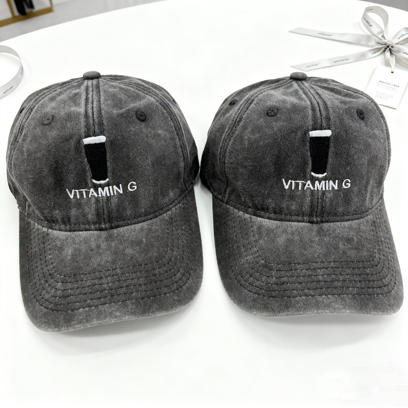 May include: Two gray baseball caps with a distressed finish. Each cap features a black graphic of a glass with a white top and the text "VITAMIN G" in white. The caps have a curved brim and a button on top.