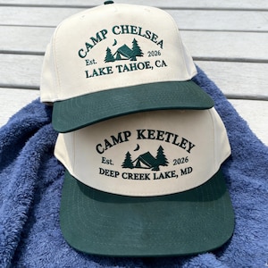 Puede incluir: Dos gorras de béisbol con corona color crema y visera verde oscuro. La gorra superior dice "CAMP CHELSEA" con "Est. 2026 LAKE TAHOE, CA" debajo de un gráfico. La gorra inferior dice "CAMP KEETLEY" con "Est. 2026 DEEP CREEK LAKE, MD".