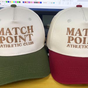 May include: Two baseball caps with the text "MATCH POINT ATHLETIC CLUB". One cap has a cream-colored crown and a green brim, while the other has a cream-colored crown and a burgundy brim. Both caps have a small button on top.