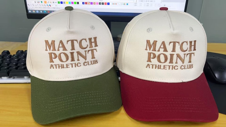 May include: Two baseball caps with the text "MATCH POINT ATHLETIC CLUB". One cap has a cream-colored crown and a green brim, while the other has a cream-colored crown and a burgundy brim. Both caps have a small button on top.