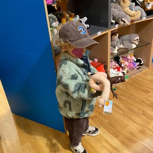 May include: A brown baseball cap with a blue and white number 3 patch, a light green and grey patterned fleece jacket, and dark brown corduroy pants. The item being held is a plush toy shaped like a pink worm attached to a brown textured branch.