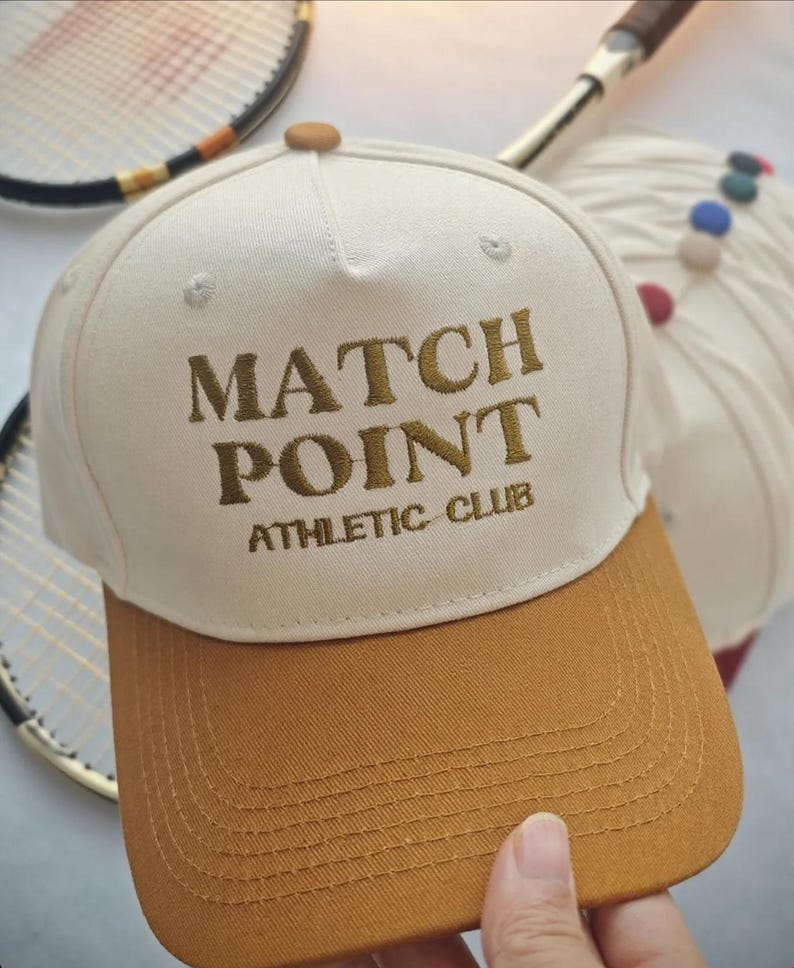 May include: A cream-colored baseball cap with a brown brim and the embroidered text "MATCH POINT ATHLETIC CLUB." The cap is a classic style, suitable for sports or casual wear.