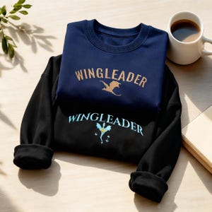 Op de afbeelding: Twee sweatshirts, één marineblauw en één zwart, zijn op elkaar gestapeld. Het marineblauwe sweatshirt heeft "WINGLEADER" in goud met een drakenafbeelding. Het zwarte sweatshirt heeft "WINGLEADER" in teal met een drakenafbeelding. Een witte koffiemok staat in de buurt.