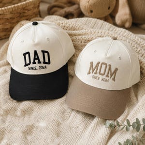 Puede incluir: Dos gorras de béisbol con coronas color blanco hueso. La gorra izquierda tiene bordado en negro DAD SINCE 2024 y visera negra. La gorra derecha tiene bordado en color canela MOM SINCE 2024 y visera color canela. Ambas tienen un botón superior.