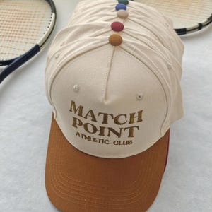 May include: A stack of beige baseball caps with brown visors. The caps have the text "MATCH POINT ATHLETIC CLUB" embroidered on the front. Each cap is adorned with a row of colorful buttons along the top. Tennis rackets are in the background.