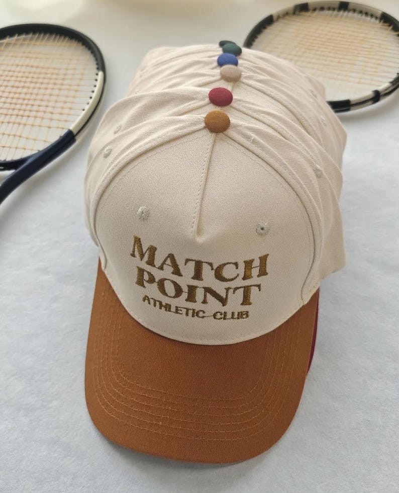 May include: A stack of beige baseball caps with brown visors. The caps have the text "MATCH POINT ATHLETIC CLUB" embroidered on the front. Each cap is adorned with a row of colorful buttons along the top. Tennis rackets are in the background.