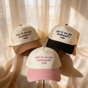 Può includere: Tre cappellini da baseball con la scritta "Put it on my brother's tab" e "Put it on my husband's tab". I cappellini hanno diverse combinazioni di colori: beige e marrone, beige e nero e beige e rosa.