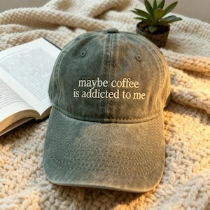 May include: A sage green baseball cap with the embroidered text "maybe coffee is addicted to me". The hat is made of a soft, worn material. A book and small potted plant are in the background.