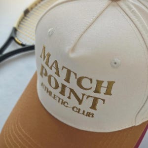 May include: Cream-colored baseball cap with a brown brim and the words "MATCH POINT ATHLETIC CLUB" embroidered in gold. The cap has a red underside and colorful button details on top. A tennis racket is visible in the background.