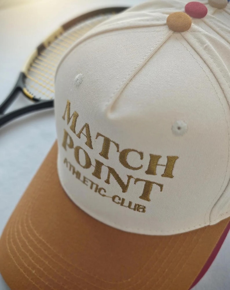 May include: Cream-colored baseball cap with a brown brim and the words "MATCH POINT ATHLETIC CLUB" embroidered in gold. The cap has a red underside and colorful button details on top. A tennis racket is visible in the background.
