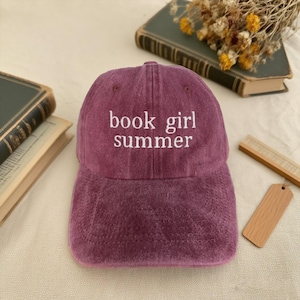 May include: A maroon baseball cap with the words "book girl summer" embroidered in white. The cap has a curved peak and a classic six-panel design. The hat is made of a soft, textured fabric.