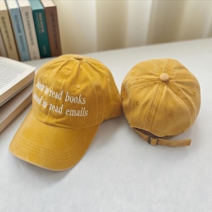 May include: Two mustard-yellow baseball caps. One cap has the embroidered text "Born to read books forced to read emails" in white. The other cap is shown from the back, with an adjustable strap. The caps are on a white surface.
