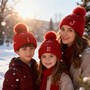 May include: Three people wearing matching red knit hats with white embroidered letters and a fluffy pom-pom. The hats are paired with red scarves and jackets. The scene is set outdoors in a snowy environment, suggesting a winter theme.