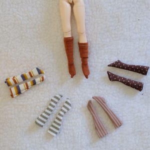 May include: A collection of doll socks in various patterns and colors. Includes pairs of striped socks in blue, yellow, and white, gray and white striped socks, brown socks with white polka dots, and solid brown and pink socks. Doll legs are also visible.