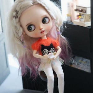 May include: A doll with long, wavy, light-colored hair with pink and purple highlights. The doll wears an orange shirt with a black cat design, a white skirt, and white leggings. It holds a small white teacup and saucer.
