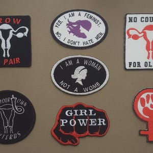 Peut inclure: Collection de patchs brodés avec des slogans féministes et des graphiques d'utérus. Les patchs présentent des phrases telles que « Girl Power », « I am a woman, not a womb » et « No country for old man ». Les couleurs incluent le noir, le blanc, le rouge et le violet.