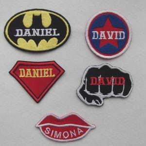 May include: A collection of embroidered name patches in various shapes. Designs include a Batman logo, a star in a circle, a diamond, a fist, and a pair of lips. Each patch has a name in white lettering against a coloured background. The patches are in black, red, yellow, and blue.