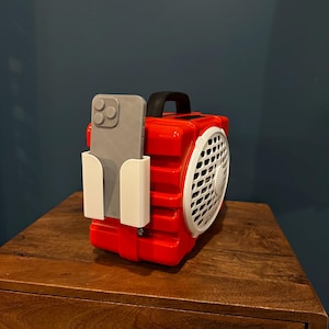 May include: A red and white portable fan with a black handle and a white phone holder. A gray smartphone is placed in the holder. The fan is on a wooden bedside table.
