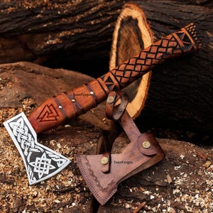 May include: A handcrafted axe with a wooden handle featuring intricate carvings and leather accents. The axe head is silver with a Celtic knot design. A leather sheath protects the blade. The axe is displayed on a bed of wood shavings.