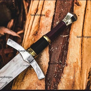 May include: A medieval sword with a silver blade and crossguard, a black handle, and a burgundy grip. The sword rests on a wooden surface. The text "SvenForges" is visible on the image.
