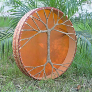 Pode incluir: Um tambor redondo feito à mão com um desenho de galhos de árvore. O tambor apresenta uma pele de tom laranja quente esticada sobre uma moldura de madeira. A moldura é decorada com uma borda estampada. O tambor está sobre a grama verde.