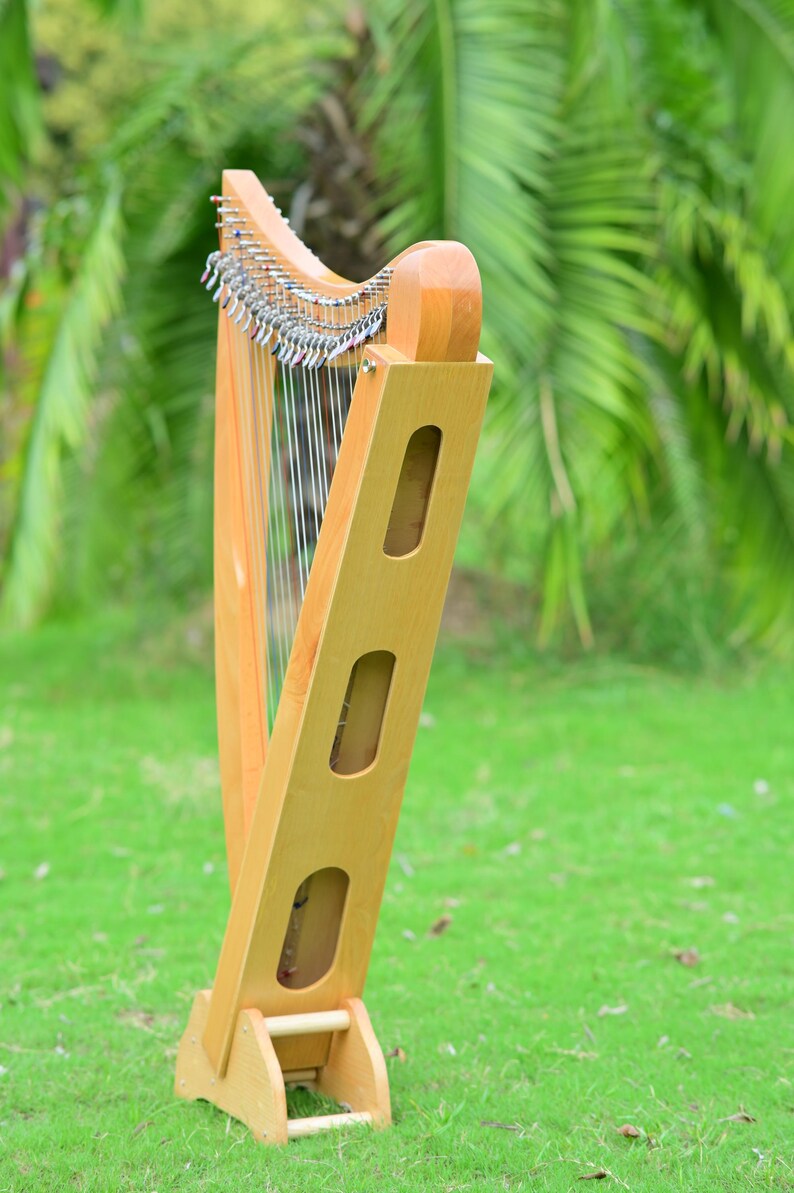May include: A wooden harp with a light brown finish stands on green grass. The harp has a curved top with metal tuning pegs and numerous strings. The body features three elongated cutouts. The background is blurred with green foliage.