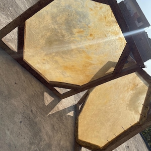 May include: Two octagonal drum frames with stretched, light-colored hides. The wooden frames are constructed with angled supports. The hides have a natural, textured appearance, with variations in color. The drums are positioned on a concrete surface.
