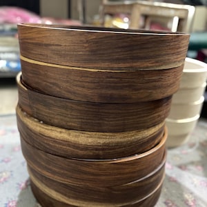 Puede incluir: Una pila de seis cuencos de madera marrón oscuro con un patrón de vetas de madera natural. Cada cuenco tiene una fina banda de color más claro cerca de la parte superior. Los cuencos están apilados, con algunos cuencos blancos visibles en el fondo.