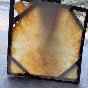 May include: A square, tanned hide stretched within a dark metal frame. The hide is a light tan color with darker brown areas, and it has a slightly translucent appearance. The frame has a geometric design.