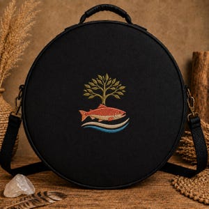 May include: A round black crossbody bag featuring an embroidered design of a green tree above a salmon fish swimming over stylized blue and white waves. The bag has a top handle, a black adjustable strap with metal clips, and a zipper closure.