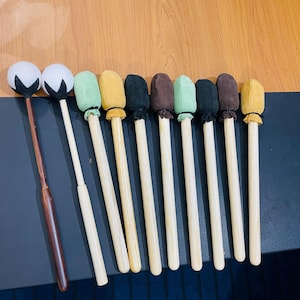 Op de afbeelding: Een verzameling percussiehamers met verschillende gekleurde koppen, waaronder wit, groen, geel, zwart en bruin. De hamers hebben houten handvatten, waarvan één hamer een donkerder, roodbruin handvat heeft. De hamers zijn gerangschikt op een donkere ondergrond.