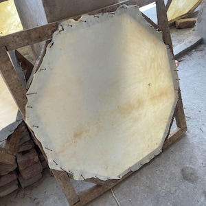 May include: A large, irregularly shaped piece of off-white leather stretched and secured to a wooden frame with small nails. The leather has a slightly textured surface and some areas of discoloration. The frame is made of rough-hewn wood.
