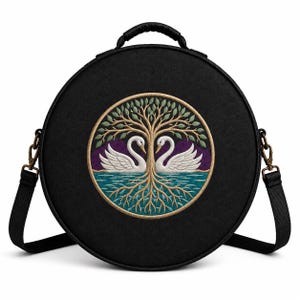 May include: A round black crossbody bag featuring a central circular embroidery of two white swans facing each other beneath a tree with green leaves and visible roots. The bag includes a top handle and an adjustable black shoulder strap with metal clips.
