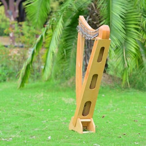 May include: A wooden harp with a light brown finish stands on green grass. The harp has a curved top with strings and a rectangular frame with three oval cutouts. The background features green foliage and a blurred view of a structure.