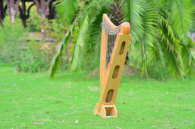 May include: A wooden harp with a light brown finish stands on green grass. The harp has a curved top with strings and a rectangular frame with three oval cutouts. The background features green foliage and a blurred view of a structure.