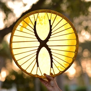 May include: A circular, yellow drum with a dark brown, symmetrical design in the centre. The drum is held up against a blurred background of trees and sunlight. The drum's frame is a light brown colour.