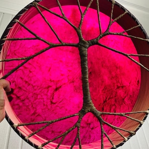 May include: A round, decorative piece featuring a vibrant pink center and a tree-like design. The tree branches extend across the pink surface. The outer rim is a dark brown, and the piece is held by a hand. The overall design is artistic and eye-catching.