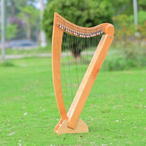 May include: A wooden harp with a light brown finish stands on a grassy lawn. The instrument has a curved neck and a triangular body, with numerous strings stretched across its frame. The harp is designed to be played and is a musical instrument.