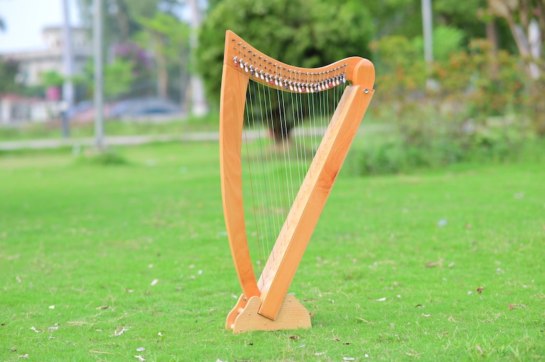 May include: A wooden harp with a light brown finish stands on a grassy lawn. The instrument has a curved neck and a triangular body, with numerous strings stretched across its frame. The harp is designed to be played and is a musical instrument.