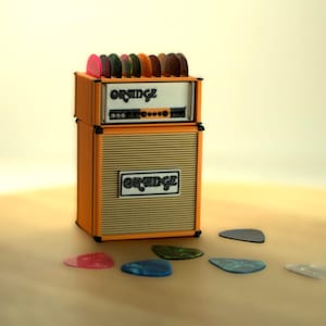 May include: A miniature orange guitar amplifier, branded with "ORANGE", with a collection of guitar picks on top and scattered in front. The amp is a replica, with a speaker grille and control panel details.