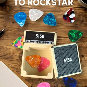 Pode incluir: Uma superf&iacute;cie de madeira exibe uma cole&ccedil;&atilde;o de palhetas de guitarra em v&aacute;rias cores e padr&otilde;es, incluindo azul, branco e vermelho. Um pequeno amplificador com o n&uacute;mero "5150" &eacute; apresentado, juntamente com o texto "FROM CLUTTER TO ROCKSTAR" e "Magnetic half stack".