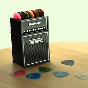 May include: A small black guitar amplifier, branded with the "Blackstar" logo, and a selection of guitar picks in various colours. Several guitar picks are scattered on the wooden surface in front of the amp. The amp has a speaker grill and control knobs.