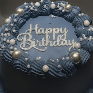 Peut inclure: Un gâteau bleu givré avec les mots "Happy Birthday" en écriture argentée. Le gâteau est décoré de tourbillons de glaçage bleu, de perles blanches et de sphères métalliques argentées. Un dessert de fête.