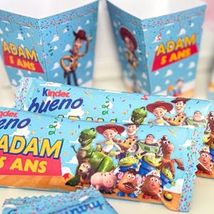 May include: Blue party boxes and chocolate bars with Toy Story characters. The boxes display "ADAM 5 ANS" in red and yellow. The chocolate bars are branded "Kinder Bueno". Confetti and clouds decorate the background.