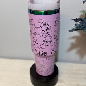 May include: A pink tumbler with a white lid and a black base. The tumbler is covered in signatures and character names in black ink. The top rim has a green and iridescent band. The tumbler is standing on a black, round base.