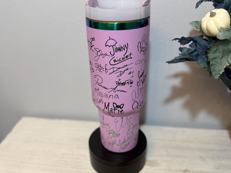 May include: A pink tumbler with a white lid and a black base. The tumbler is covered in signatures and character names in black ink. The top rim has a green and iridescent band. The tumbler is standing on a black, round base.